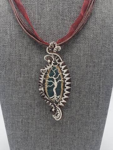 Ornate silver pendant on a burgundy ribbon necklace displayed on a gray jewelry bust.