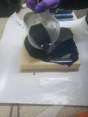 Person in purple gloves pouring dark liquid onto light base for acrylic string pull soap art