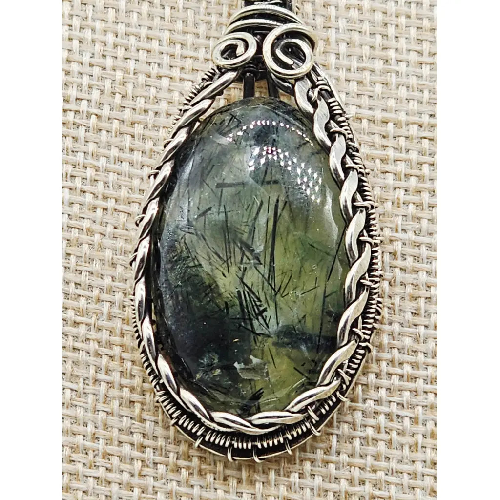 Oval tourmalinated prehnite pendant in sterling silver wire wrapping. Front view of a pale green gemstone with black tourmalinated inclusions.